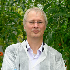 A person with glasses and light hair in a white collared shirt and protective gear stands before lush green plants, showcasing Fluence's LED lighting for optimized controlled environment agriculture, including cannabis cultivation, Austin, TX.