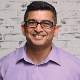 Smiling man in glasses and purple shirt, expert in LED grow lights for cannabis, white brick backdrop.