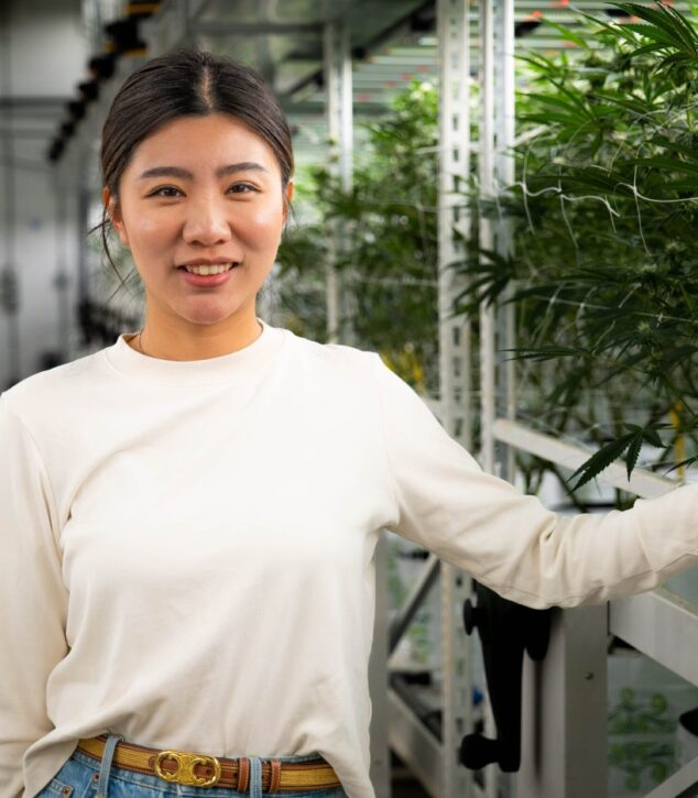 Smiling person in greenhouse touching LED lighting structure by Fluence; vibrant green plants in background, Austin-based controlled environment agriculture lighting experts specializing in crop yield optimization including cannabis.