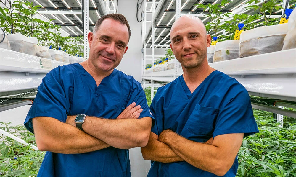 Two growers in scrubs stand under LED grow lights in a cannabis cultivation facility.