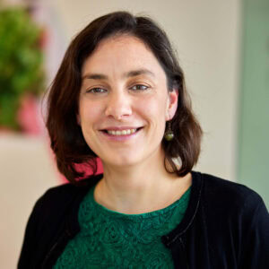 A smiling woman with shoulder-length brown hair in a green shirt and black cardigan, representing Fluence LED lighting solutions for cannabis cultivation, stands indoors against a blurred backdrop, highlighting optimized crop production in controlled environments. width=
